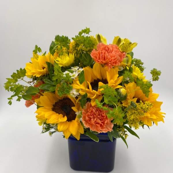 Low arrangement of yellow sunflowers and orange carnations in a blue cube vase