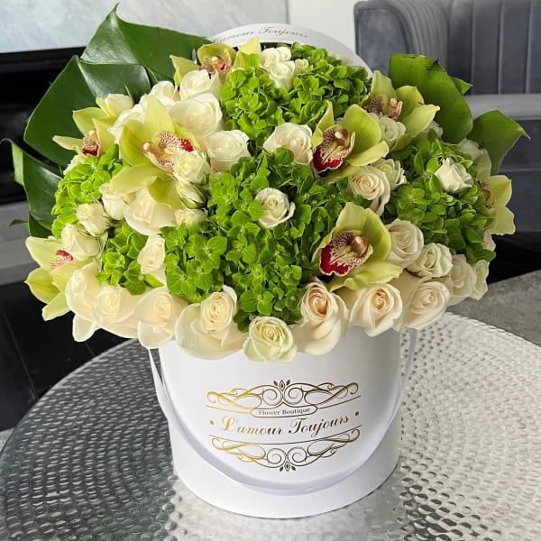 Round box arrangement of cream roses, green hydrangeas, and green orchids on a glass table