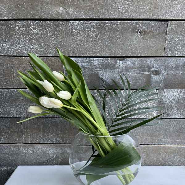 White tulips arranged in a round glass vase with palm leaves