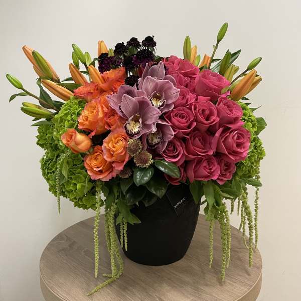 Lush arrangement of bright pink roses, orange lilies and roses, orchids and green hydrangeas in a black pot.