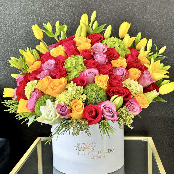 Bright arrangement of multicolor roses, yellow tulips, lilies, and green blooms in a white hat box.