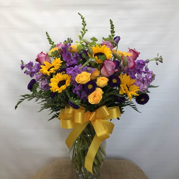 Bouquet of sunflowers, purple flowers, and pink roses in a glass vase with a yellow ribbon.