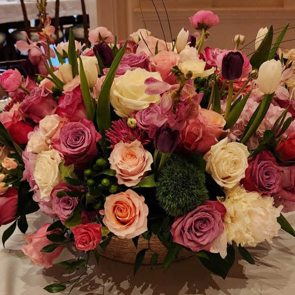 Low centerpiece of pink, purple, peach, and white roses with tulips and mixed blooms in a round bowl vase.