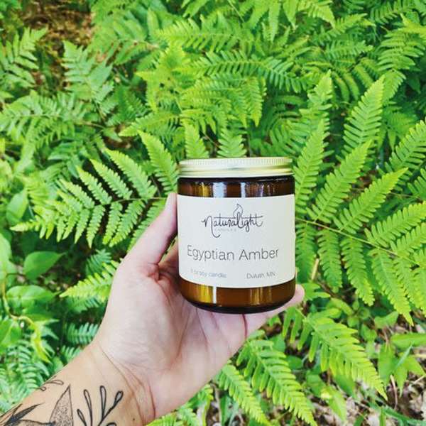 Hand holding an amber jar candle labeled Egyptian Amber in front of bright green ferns.
