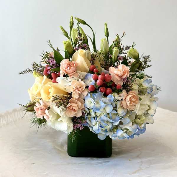 Low mixed arrangement of peach carnations, yellow roses and blue hydrangeas in a dark green glass cube vase