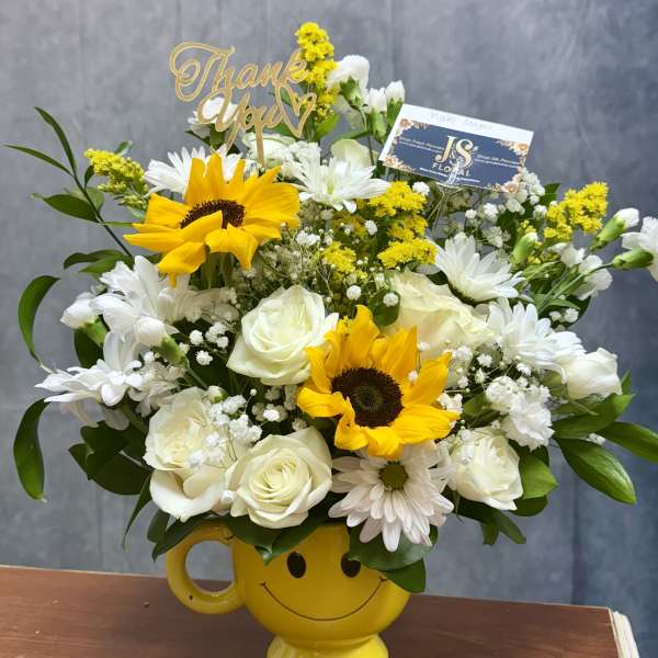 Cheerful bouquet of sunflowers, white roses, and daisies in a yellow smiley mug with a thank you pick.