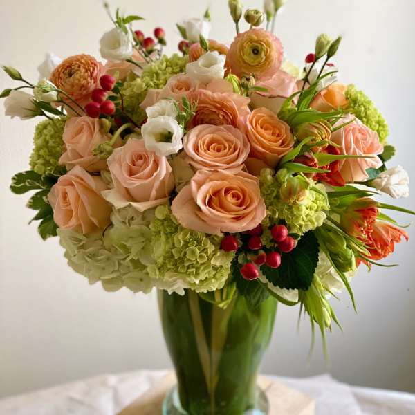 Tall vase arrangement of peach roses, green hydrangeas, white blooms and red berries.