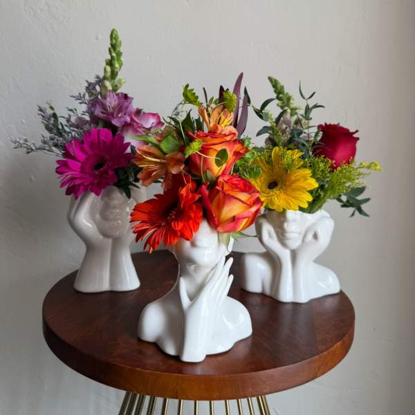 Three colorful flower arrangements in white face-shaped vases on a round table