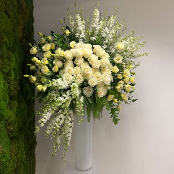Tall grand arrangement of ivory roses and white flowers in a white pedestal vase against a moss wall