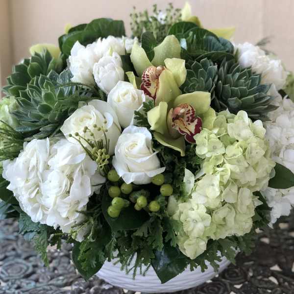 Low round arrangement of white roses, hydrangeas, green succulents, and orchids in a white bowl