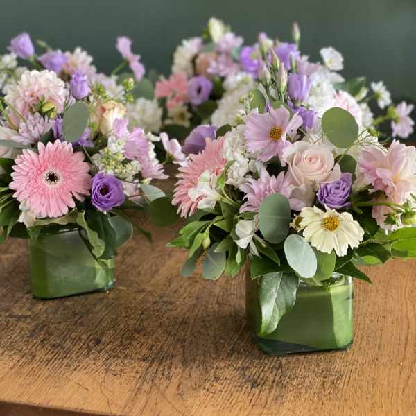 Two pastel floral arrangements in square glass vases on a wooden table