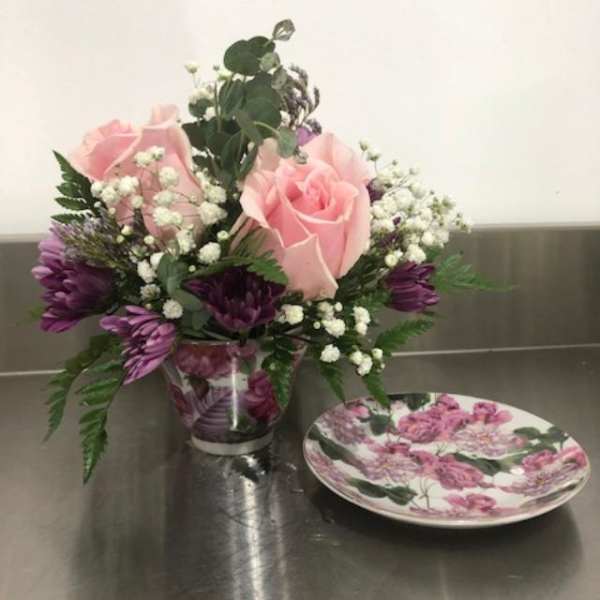 Pink roses and purple flowers in a clear vase beside a floral plate