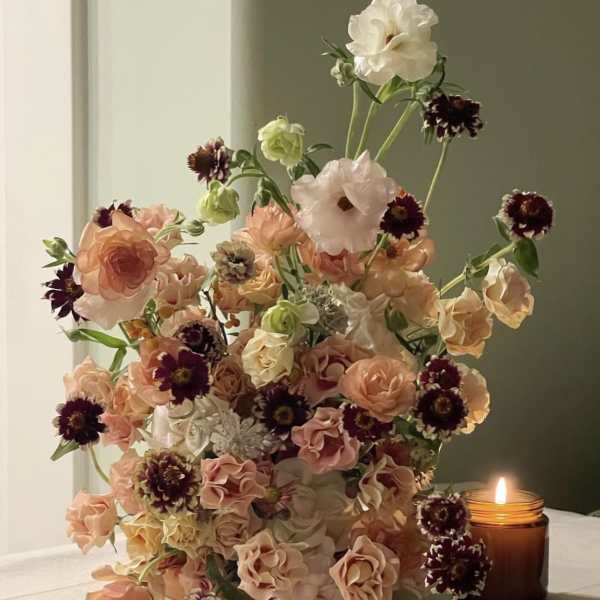 Tall bouquet of peach, blush, white, and burgundy flowers beside a lit candle