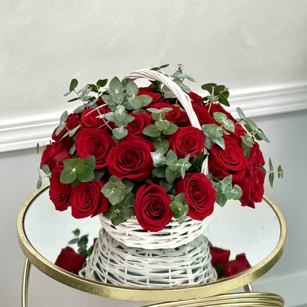 Round basket of red roses with eucalyptus in a white woven handle basket on a mirrored table