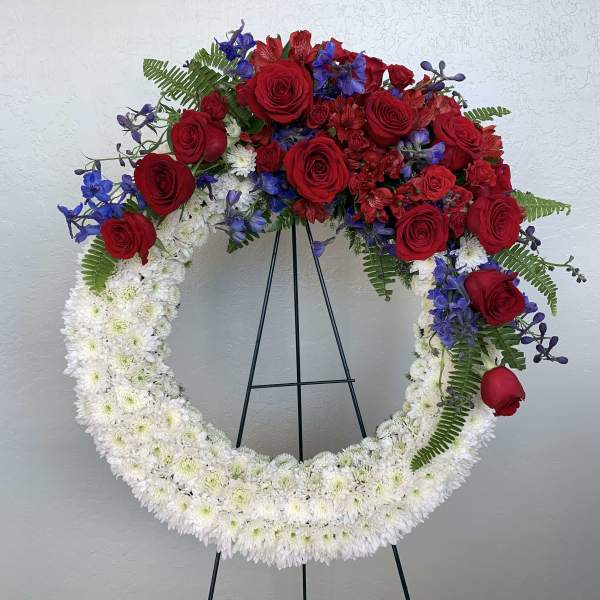 Red roses and blue flowers arranged in a large white floral wreath on a stand.