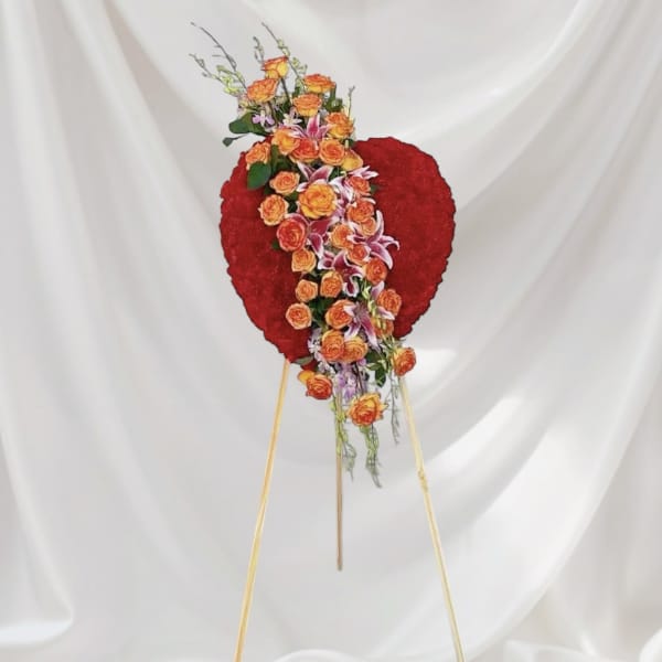 Standing floral spray with orange roses and lilies on a red heart-shaped backing