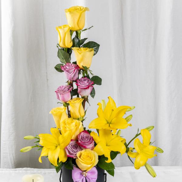 Tall arrangement of yellow lilies and yellow and lavender roses in a black box with a bow