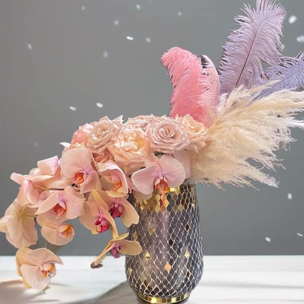 Pink orchids and roses in a patterned vase with pastel feathers