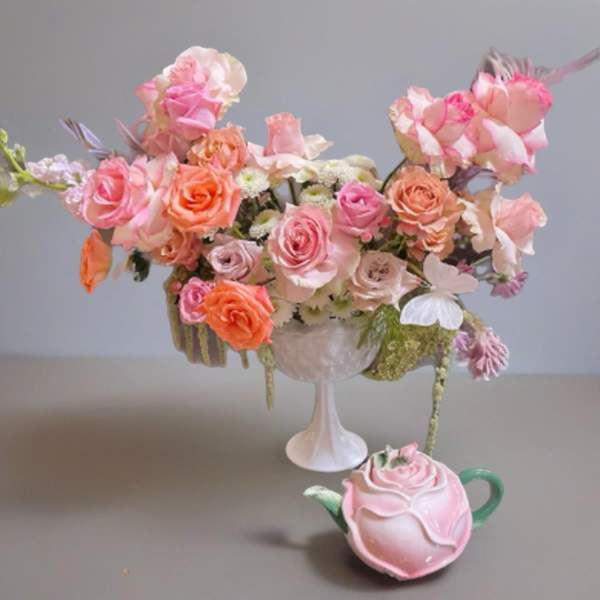 Pink and peach rose arrangement in a white pedestal vase with a rose-shaped teapot