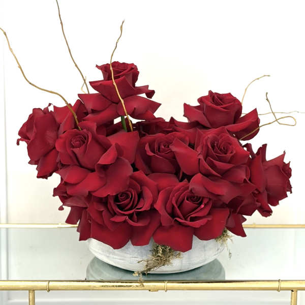 Low white bowl filled with clustered deep red roses and curly decorative branches
