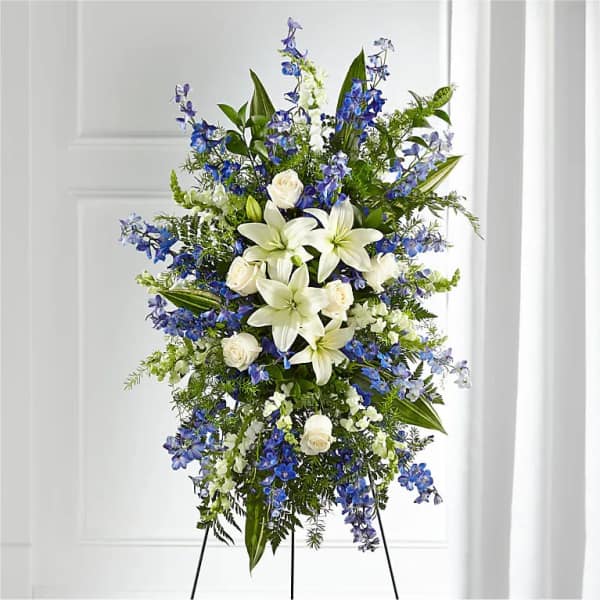 Standing spray of white lilies and roses with blue delphinium on a metal easel.