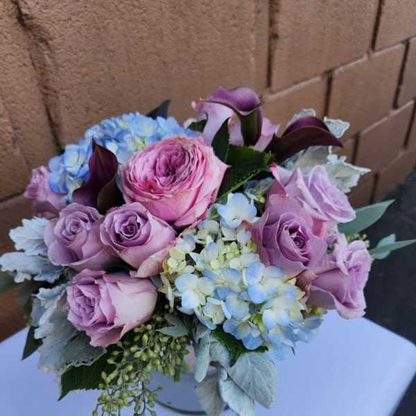 Low vase arrangement of pink and lavender roses, blue hydrangeas, and dark purple calla lilies.