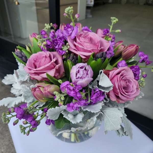 Compact arrangement of pink roses and purple flowers in a round glass vase