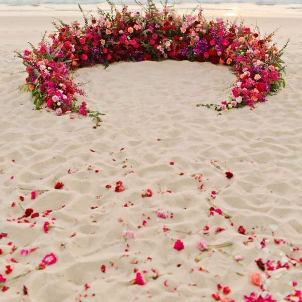 Large crescent-shaped floral arrangement on a beach at sunset