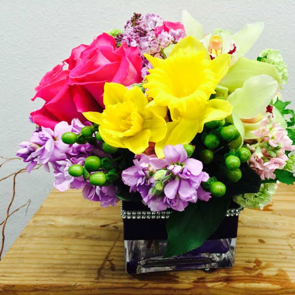 Bright mixed bouquet with pink roses, yellow daffodils, and purple flowers in a square vase