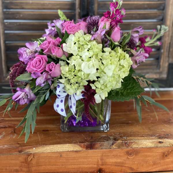 Pink roses and purple flowers in a square glass vase with a polka-dot ribbon.