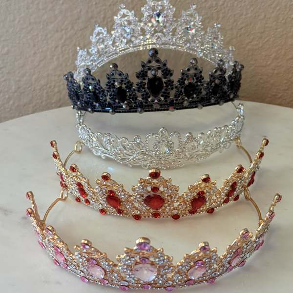 Four jeweled tiaras in silver, black, red, and pink sit stacked on a table.