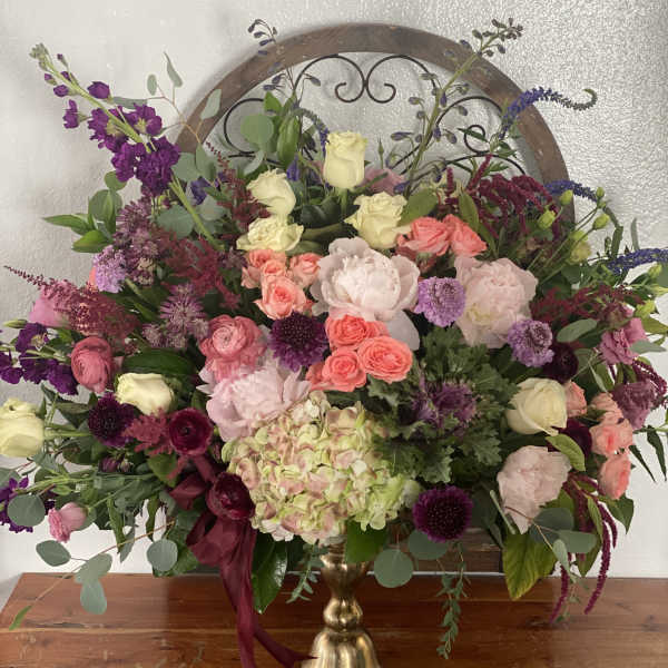 Large mixed floral arrangement in a gold vase with pink, purple, and cream blooms