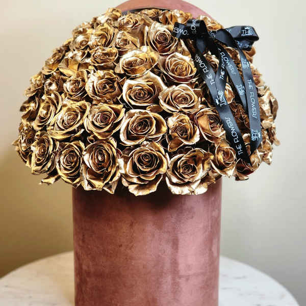 Gold roses arranged in a pink cylindrical box with a black ribbon