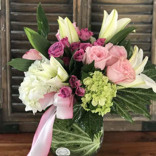 Pink and white lilies, roses, and hydrangeas in a clear glass vase with a pink ribbon bow