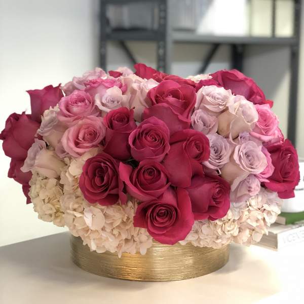 Low round arrangement of hot pink and pale pink roses with ivory hydrangeas in a gold cylinder vase