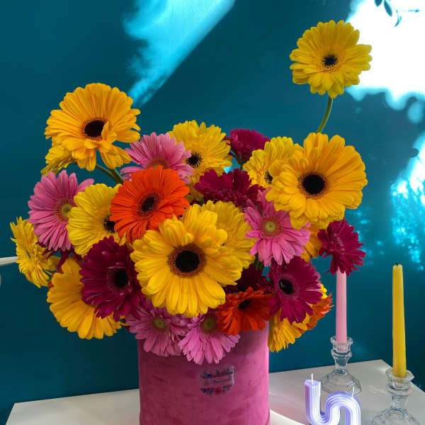 Bright gerbera daisy arrangement in a pink hatbox with candles nearby