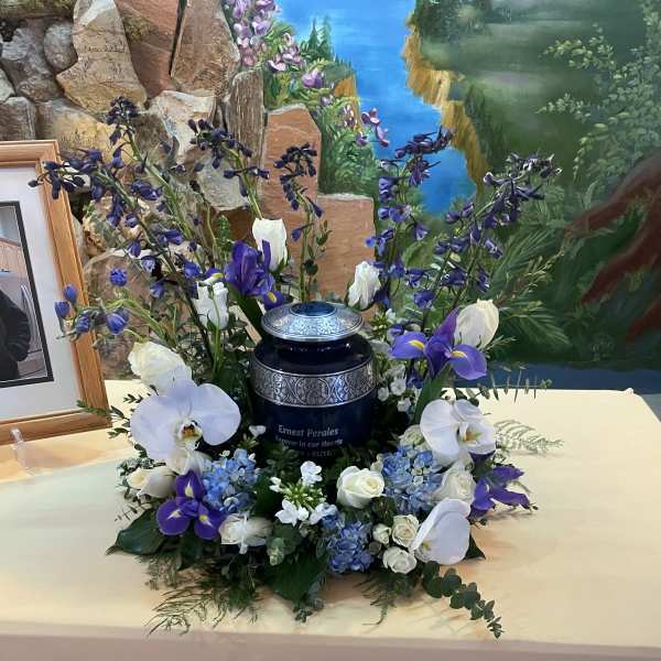 Cremation urn surrounded by blue hydrangeas, white roses, orchids and purple flowers on a draped table