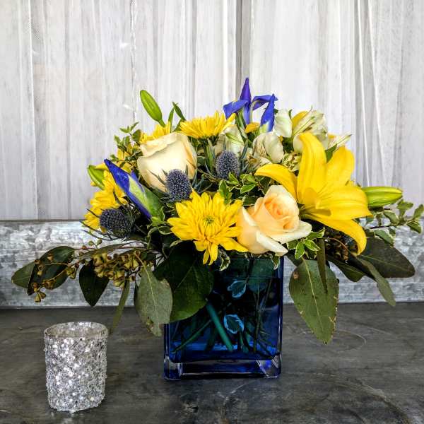 Compact arrangement of yellow lilies, cream roses, and blue accents in a blue glass cube vase.
