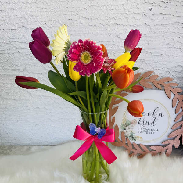 Colorful tulip and gerbera bouquet in a clear glass vase with a pink ribbon