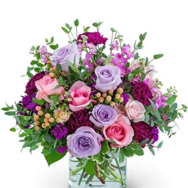 Round arrangement of pink and lavender roses with purple flowers in a clear glass vase