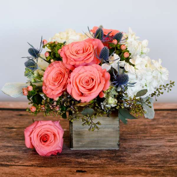 Low arrangement of coral roses, white blooms, and blue thistles in a wooden cube vase with one rose beside it