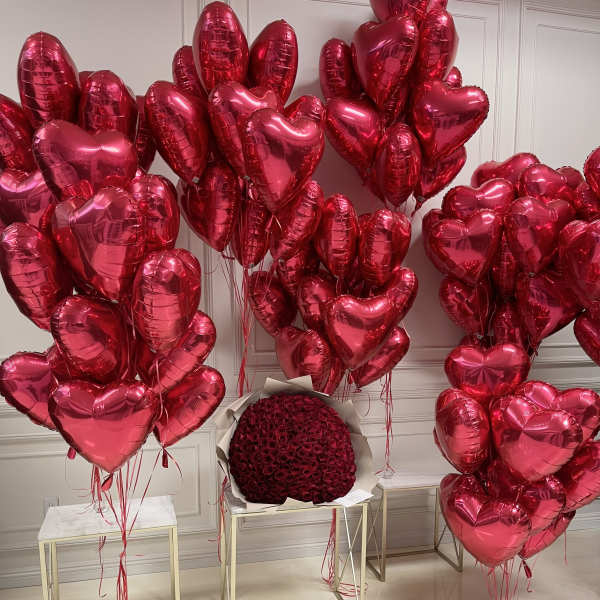 Clusters of red heart-shaped balloons surround a round bouquet of red roses.