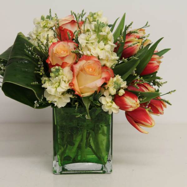 Compact arrangement of orange roses and tulips with white flowers in a green glass cube vase