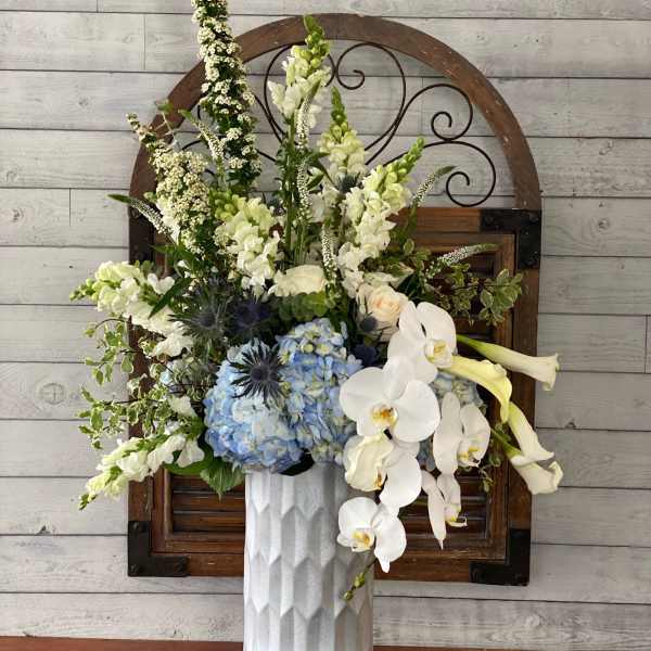 Tall floral arrangement with white orchids, blue hydrangeas, and pale blooms in a white vase