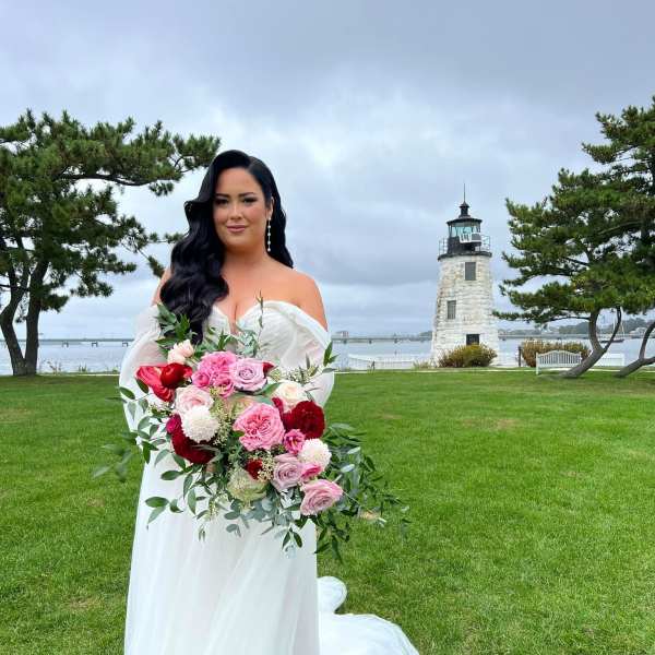 Bride holding a pink and red bouquet outdoors near a lighthouse