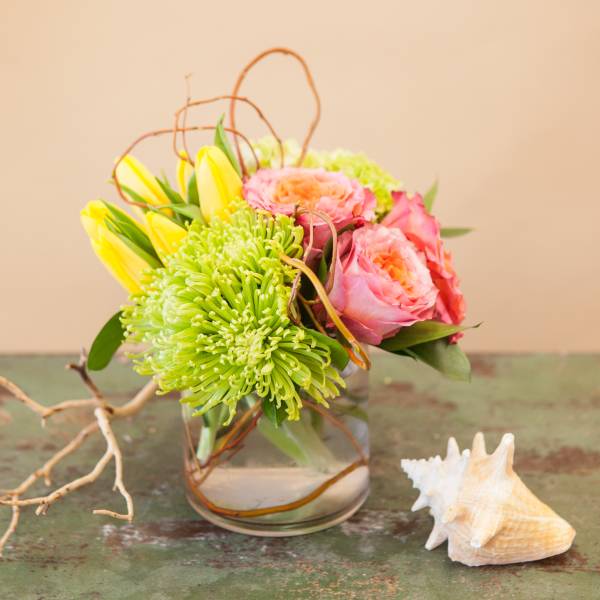 Pink and yellow flowers arranged in a clear glass vase with a seashell beside it