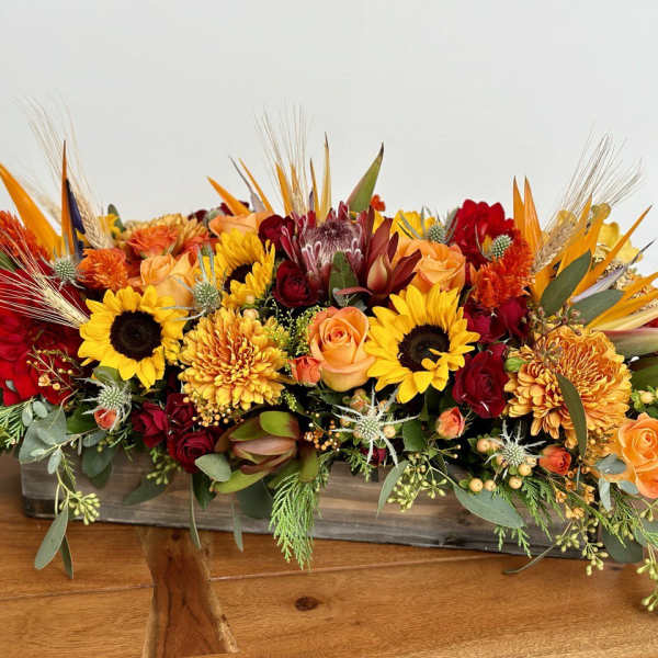 Long low arrangement of sunflowers, roses, and orange blooms in a rectangular container