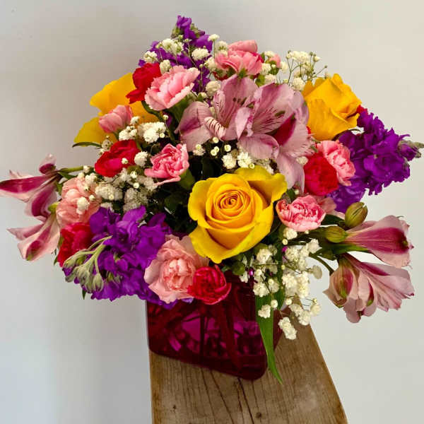 Bright mixed bouquet with yellow roses, pink carnations, and purple blooms in a pink cube vase