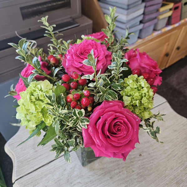 Cube vase of hot pink roses, green hydrangeas, and red berries on a light wood table