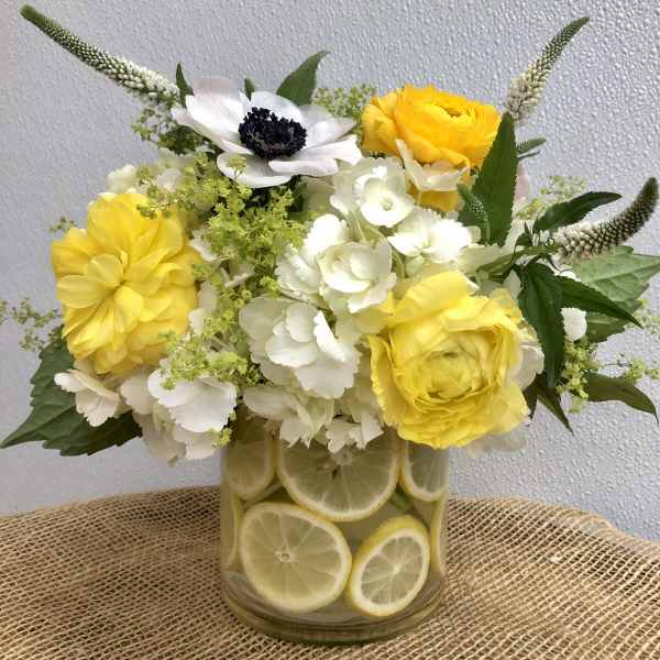 Yellow and white flower arrangement in a glass vase lined with lemon slices on a burlap surface.